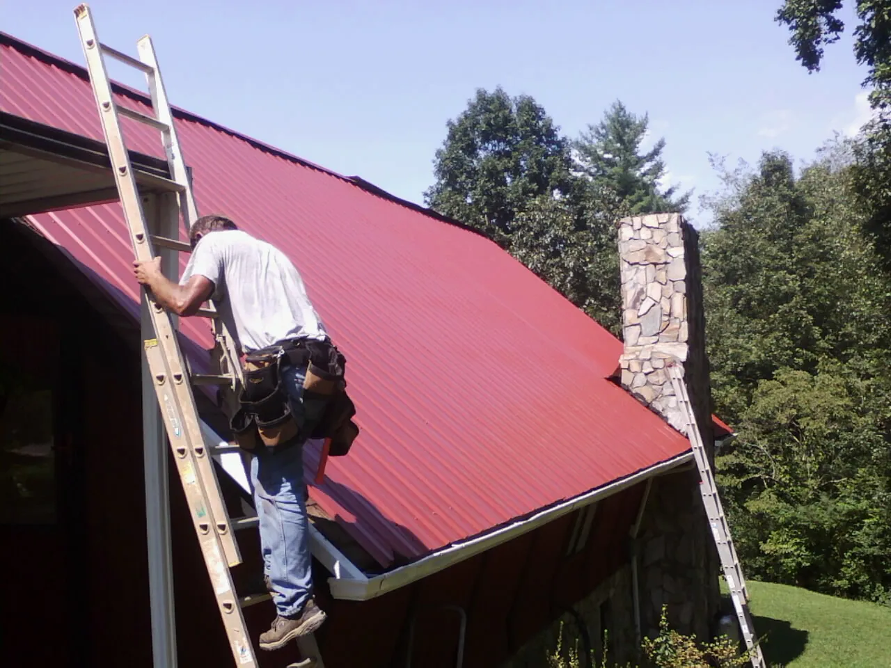Roofer on a ladder beside a red metal roof and stone chimney — Soffit & Fascia Repair by Roofing Brothers in South Seaville